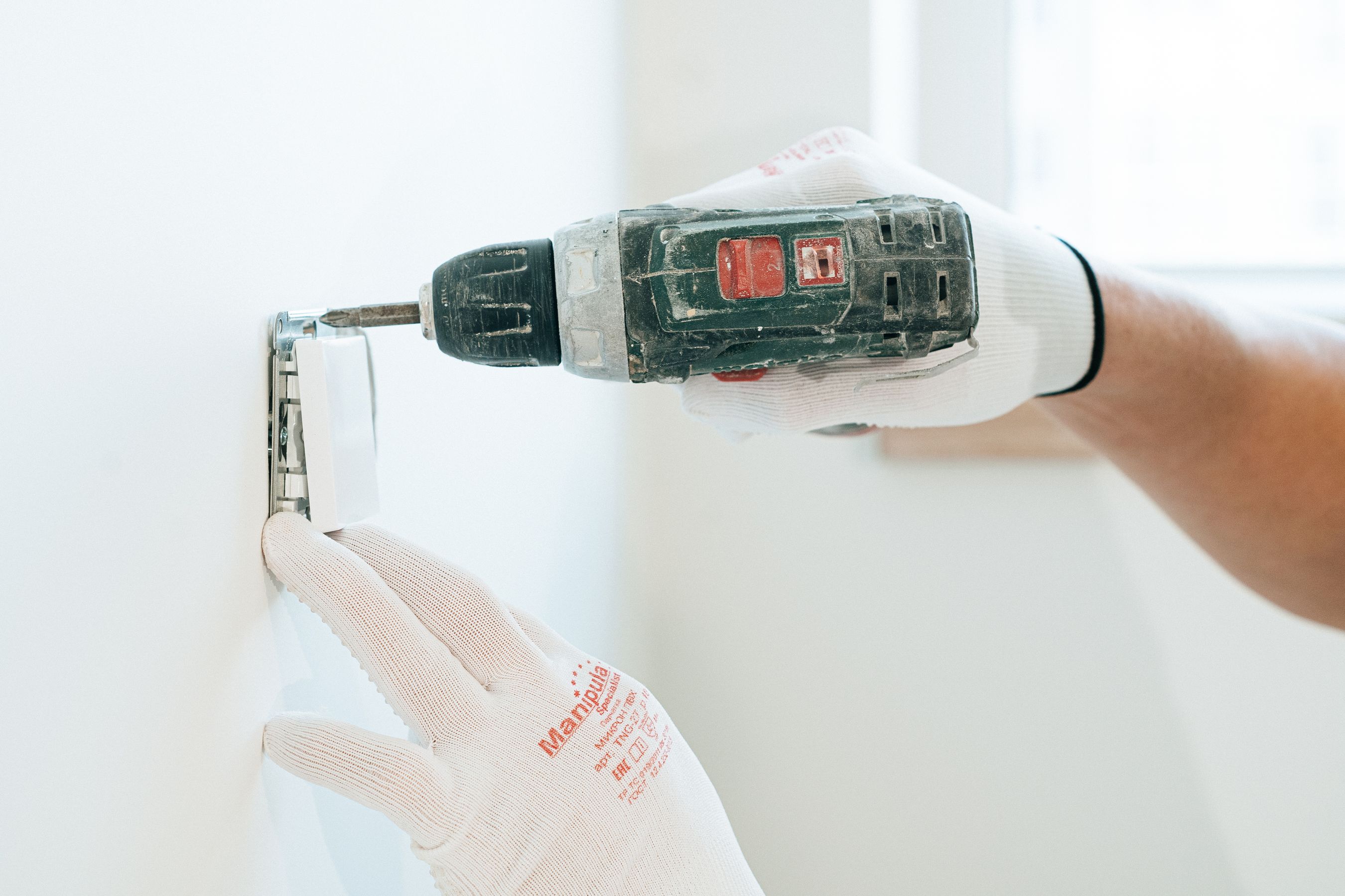 Gloved hands using a drill to install a power outlet on a wall